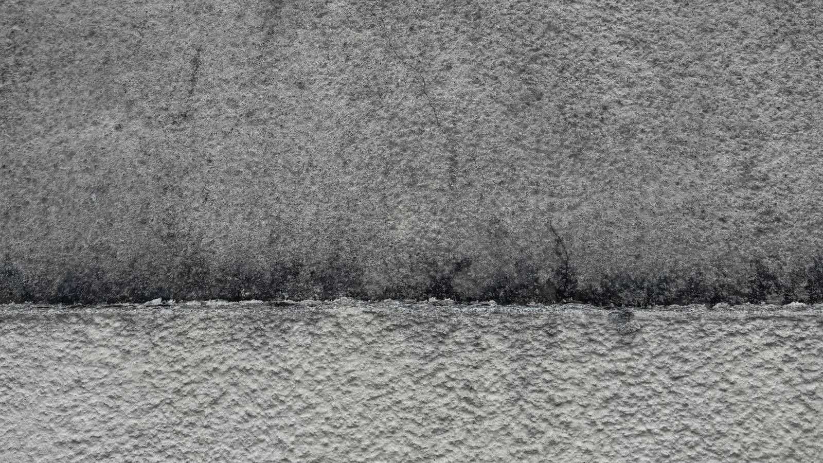 Mur en crépi gris : partie haute encrassée et microfissurée, partie basse nettoyée, la limite entre les deux est nette