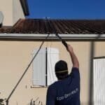 Opérateur Eco Drones de dos, perche télescopique tendue vers une toiture en tuiles canal d'une maison en crépi clair, technique adaptée à chaque chantier