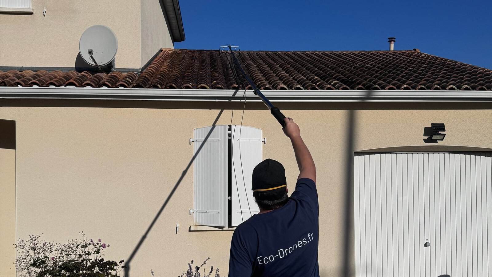 Op&eacute;rateur Eco Drones de dos, perche t&eacute;lescopique tendue vers une toiture en tuiles canal d'une maison en cr&eacute;pi clair, technique adapt&eacute;e &agrave; chaque chantier